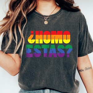 May include: Dark gray t-shirt with the text "¿COMO ESTAS?" in rainbow colors. The shirt is a casual style, suitable for everyday wear. The design features a question in Spanish, making it a unique statement piece.