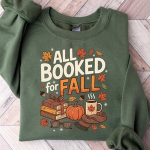 May include: Green sweatshirt with the text "ALL BOOKED for FALL" in white and orange. The design includes a stack of books, a pumpkin, a mug of coffee, and fall leaves. The sweatshirt has a crew neck and long sleeves.
