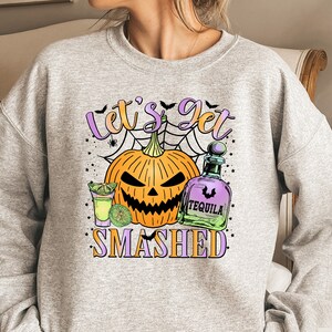 Puede incluir: Una sudadera gris con el texto "Let's Get Smashed" en letras moradas y negras. El diseño presenta una calabaza de Halloween, una botella de tequila y un vaso de chupito con una rodaja de lima.