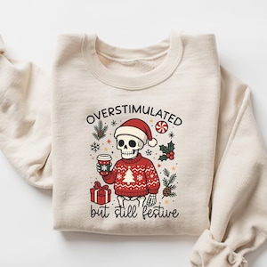Puede incluir: Sudadera color crema con un gráfico de un esqueleto con gorro de Papá Noel y un suéter navideño rojo, sosteniendo una taza de café. El texto dice "OVERSTIMULATED but still festive". Ropa festiva navideña.
