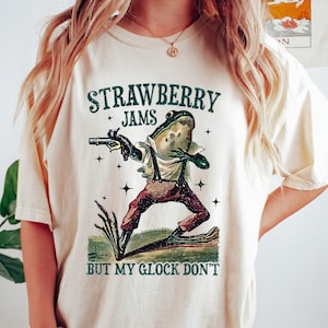 Könnte beinhalten: Cremefarbenes T-Shirt mit einem Frosch, der eine Pistole hält und Hosenträger und Hosen trägt. Der Text "STRAWBERRY JAMS" und "BUT MY GLOCK DON'T" ist über und unter der Froschillustration gedruckt. Teil einer Bekleidungskollektion.