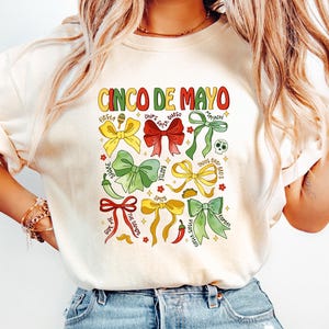 May include: A white t-shirt with the text "Cinco de Mayo" printed in black. The shirt features a colorful design of bows with various words and phrases related to the holiday, such as "Fiesta", "Tacos", and "Hot Sauce".