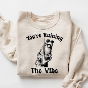 Puede incluir: Sudadera color crema con el texto "You're Ruining The Vibe" sobre una ilustración en blanco y negro de una rana con gafas de sol y sosteniendo una barra. Las mangas están enrolladas.