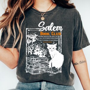 May include: Dark gray t-shirt with a white graphic featuring the text "Salem Book Club." The design includes a witch on a broom, a black cat, and a stack of books. The shirt also has the text "Books, Booze, Black Cats, All Witches Welcome."