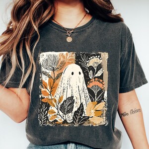 Op de afbeelding: Donkergrijs T-shirt met een vierkante afbeelding van een spook omringd door herfstbladeren en bloemen. Het spook is wit met zwarte ogen. Het T-shirt wordt gedragen met een blauwe jeans en witte schoenen. De tekst "Cozy Gifts Company" staat onderaan.