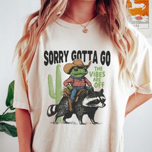 Può includere: T-shirt color crema con la scritta "SORRY GOTTA GO THE VIBES ARE OFF". La grafica presenta una rana con cappello da cowboy in sella a un procione, con un cactus. T-shirt in stile casual e unisex.