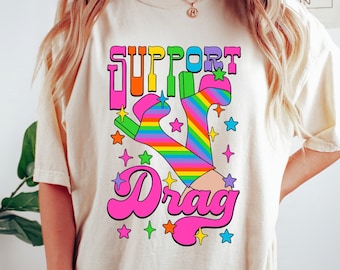May include: A white t-shirt featuring a rainbow-coloured graphic of a pair of platform boots and the text "SUPPORT Drag" in pink, yellow, and blue.