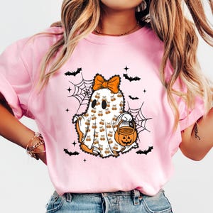 Puede incluir: Camiseta rosa claro con un dise&ntilde;o de fantasma de Halloween. El fantasma tiene un lazo naranja, lleva un cubo de calabaza y est&aacute; rodeado de murci&eacute;lagos y telara&ntilde;as. El texto "Cozy Gifts Company" est&aacute; en la parte inferior.