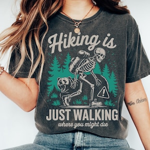 May include: Dark gray t-shirt with the text "Hiking is Just Walking Where You Might Die." The graphic features a skeleton hiker with a backpack, a bear, and a warning sign, set against a backdrop of green trees. A humorous design for outdoor enthusiasts.