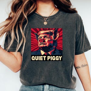 May include: Dark gray t-shirt featuring a graphic of a person's face morphed into a pig, set against a red and white background. The text "QUIET PIGGY" is displayed below the image. The shirt is worn by a person.