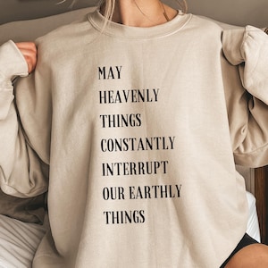 Puede incluir: Una sudadera beige con el texto "May Heavenly Things Constantly Interrupt Our Earthly Things" impreso en la parte delantera.