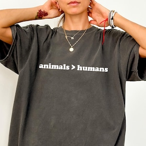 Könnte beinhalten: Dunkelgraues T-Shirt mit dem weißen Text "animals > humans". Das Shirt hat kurze Ärmel und eine lockere Passform. Das Kleidungsstück ist einfarbig und hat einen Vintage-Look.