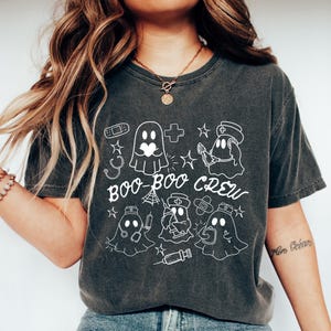 May include: Dark gray t-shirt with a white graphic of ghost characters in medical attire, including a nurse, doctor, and bandaged ghost. The text "BOO-BOO CREW" is written in a playful font. The shirt is paired with blue jeans and white sneakers.