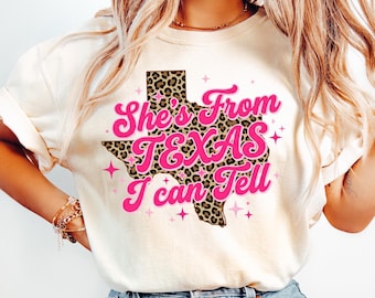 Könnte beinhalten: Cremefarbenes T-Shirt mit dem Text "She's From Texas I Can Tell" in Pink, mit einem Leopardenmuster-Texas-Umriss. Das Shirt wird mit hellblauen Jeans kombiniert.