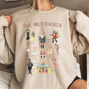 Puede incluir: Una sudadera beige con un estampado gráfico de personajes del ballet El Cascanueces. El estampado incluye a Clara, el Cascanueces, el Rey Ratón y otros personajes. El texto "THE NUTCRACKER" está impreso en marrón sobre el gráfico.