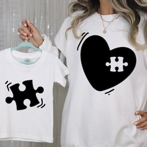 以下が含まれることがあります： 黒いパズルピースのデザインが施された、白いTシャツとスウェットシャツのセット。スウェットシャツには、パズルピースの切り抜きが付いた大きな黒いハートがあります。Tシャツには、単一のパズルピースグラフィックがあります。お揃いのセット。