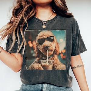 May include: Dark grey t-shirt featuring a graphic of a dog wearing sunglasses and drinking from a cup with a straw. The dog is a light brown color. The shirt is paired with blue jeans and white sneakers.