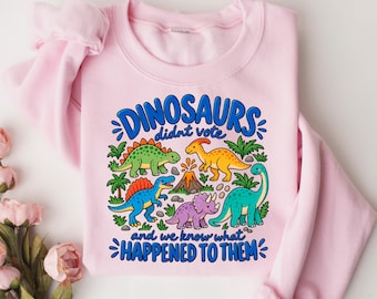 May include: A light pink crewneck sweatshirt featuring a colorful graphic of five dinosaurs, palm trees, and a volcano. The text reads "DINOSAURS didn't vote AND WE KNOW WHAT HAPPENED TO THEM" in bold blue letters. The design includes green, yellow, blue, and purple.