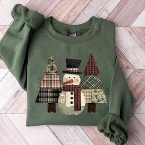 Könnte beinhalten: Ein olivgrünes Sweatshirt mit einem Schneemann und zwei auf der Vorderseite applizierten Weihnachtsbäumen. Der Schneemann hat einen schwarzen Zylinder, eine Karottennase und einen bordeauxfarbenen Schal. Die Bäume sind aus kariertem Stoff.