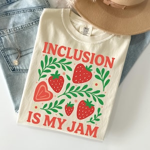 May include: Cream-colored t-shirt with the words "INCLUSION IS MY JAM" in red, along with strawberry and leaf graphics. The shirt is laid on top of blue jeans, a tan hat, and a necklace.