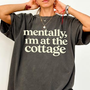 以下が含まれることがあります： 「mentally, I'm at the cottage」の白い文字がプリントされた、濃いグレーのTシャツ。半袖でゆったりとしたフィット感。カジュアルで快適なスタイルです。