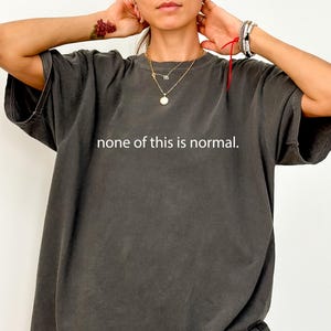 May include: Dark grey t-shirt featuring the white text "none of this is normal". The shirt has a relaxed fit and short sleeves. The fabric has a slightly faded appearance.