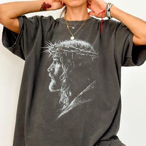 May include: Dark gray oversized t-shirt featuring a detailed white graphic of a man wearing a crown of thorns. The graphic is a side profile of the man's face and shoulders. The shirt has short sleeves and a relaxed fit.