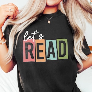 Puede incluir: Camiseta negra con el texto "let's READ" en letras de bloque de colores.