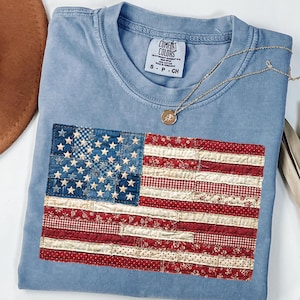 May include: A pale blue Comfort Colors t-shirt with a sewn-on American flag design. The flag features a patchwork design with red and white stripes and blue stars. A gold necklace with a pendant rests on the shirt.