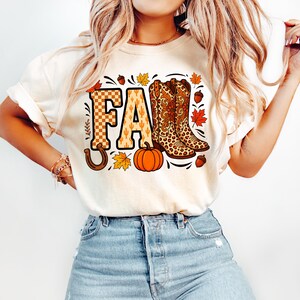 May include: A cream-colored short-sleeve t-shirt featuring a graphic print with the word FALL in large letters. The F has a checkered pattern, and the L is replaced by two leopard-print cowboy boots. The design includes a pumpkin, leaves, and acorns.