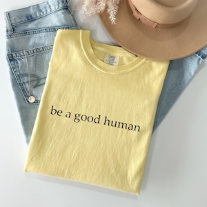 May include: A light yellow t-shirt with the phrase "be a good human" printed in black. The shirt is laid on top of light blue jeans, a tan hat, and a gold necklace.