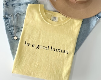 May include: A light yellow t-shirt with the phrase "be a good human" printed in black. The shirt is laid on top of light blue jeans, a tan hat, and a gold necklace.