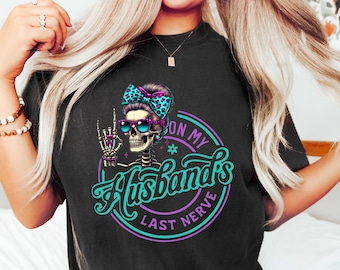 May include: Black t-shirt with a graphic of a skeleton wearing sunglasses and a bow, with the text "ON MY Husband's LAST NERVE" in a circular design. The skeleton is giving a hand gesture. The shirt is a dark color.