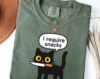 Op de afbeelding: Olijfgroen t-shirt met een illustratie van een zwarte kat met een mes en de tekst "i require snacks" in een tekstballon. Het shirt heeft een ronde hals en korte mouwen. Een gouden ketting ligt over het shirt.