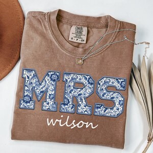 May include: Brown t-shirt with the word "MRS" in blue floral print, and the name "wilson" in white script. A silver necklace with a pendant is on the shirt. The shirt is folded on a white surface.