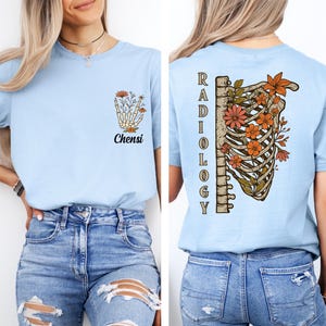 Puede incluir: Una camiseta azul claro con un pequeño gráfico en el pecho izquierdo de una mano esquelética sosteniendo flores con el texto Chensi. La parte posterior muestra una gran ilustración de una caja torácica humana llena de flores naranjas y la palabra Radiology.