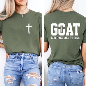 Könnte beinhalten: Olivgrünes T-Shirt mit einem weißen Kreuz auf der Vorderseite. Auf der Rückseite des Shirts steht das Wort "GOAT" mit einem Kreuz im "O" und dem Text "GOD OVER ALL THINGS."
