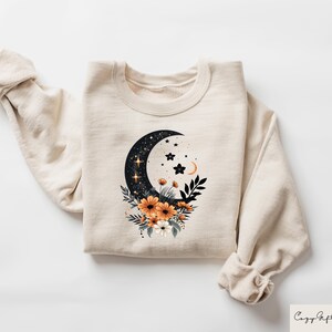 May include: Cream-colored sweatshirt featuring a crescent moon design filled with stars and a floral arrangement of orange and white flowers. The design is centered on the front of the sweatshirt. The sleeves are rolled up. The text "Cozy Gifts Company" is in the bottom right corner.