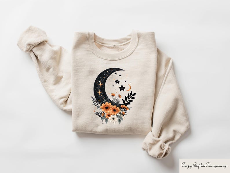 May include: Cream-colored sweatshirt featuring a crescent moon design filled with stars and a floral arrangement of orange and white flowers. The design is centered on the front of the sweatshirt. The sleeves are rolled up. The text "Cozy Gifts Company" is in the bottom right corner.