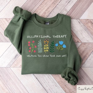 以下が含まれることがあります： 「Occupational Therapy」という文字と、「Helping you grow your own way」という文字が書かれたカラフルな花が並んだ緑色のスウェットシャツです。