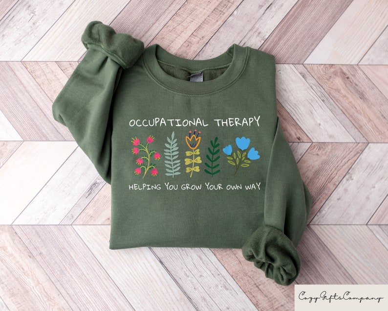 以下が含まれることがあります： 「Occupational Therapy」という文字と、「Helping you grow your own way」という文字が書かれたカラフルな花が並んだ緑色のスウェットシャツです。