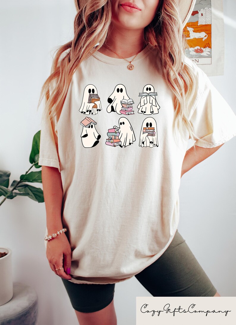 Puede incluir: Una camiseta blanca con un gr&aacute;fico de seis fantasmas sosteniendo pilas de libros. Los fantasmas son blancos con contornos negros y los libros son rosas, naranjas y marrones. La camiseta es de corte holgado y tiene cuello redondo.
