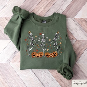 May include: A green sweatshirt with a graphic of three skeletons dancing with pumpkins and fall leaves. The skeletons are wearing white and the pumpkins are orange with black faces.