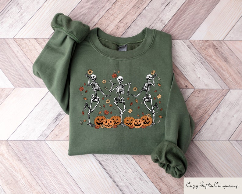 May include: A green sweatshirt with a graphic of three skeletons dancing with pumpkins and fall leaves. The skeletons are wearing white and the pumpkins are orange with black faces.