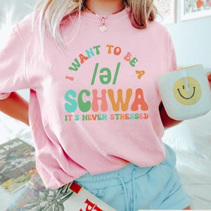 May include: Pink t-shirt with a colorful graphic that says "I want to be a /e/ SCHWA It's never stressed".