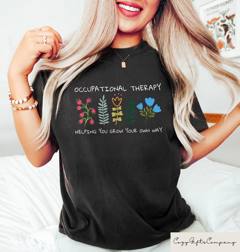以下が含まれることがあります： 「Occupational Therapy」という文字と、その下に「Helping you grow your own way」という文字が書かれたカラフルな花が並んだ黒いTシャツです。
