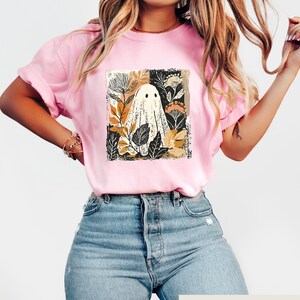 Op de afbeelding: Lichtroze T-shirt met een grillig spookontwerp omringd door botanische elementen in bruin, zwart en goud. Het shirt heeft korte mouwen en een relaxte pasvorm. De tekst "Cozy Gifts Company" staat onderaan.
