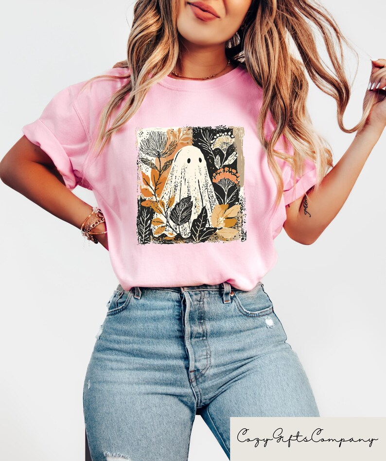 Op de afbeelding: Lichtroze T-shirt met een grillig spookontwerp omringd door botanische elementen in bruin, zwart en goud. Het shirt heeft korte mouwen en een relaxte pasvorm. De tekst "Cozy Gifts Company" staat onderaan.