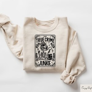 May include: A beige sweatshirt featuring a black and white graphic print of a skeleton holding a magnifying glass and a glass of wine. The graphic is in the shape of a playing card and reads "The True Crime Junkie".