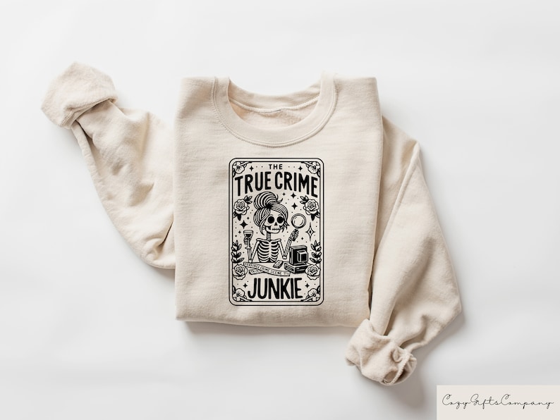 May include: A beige sweatshirt featuring a black and white graphic print of a skeleton holding a magnifying glass and a glass of wine. The graphic is in the shape of a playing card and reads "The True Crime Junkie".